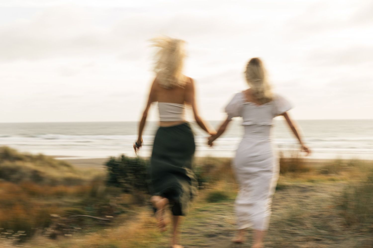 Two women holding hands walking toward the beach on a breezy day, captured with motion blur effect.