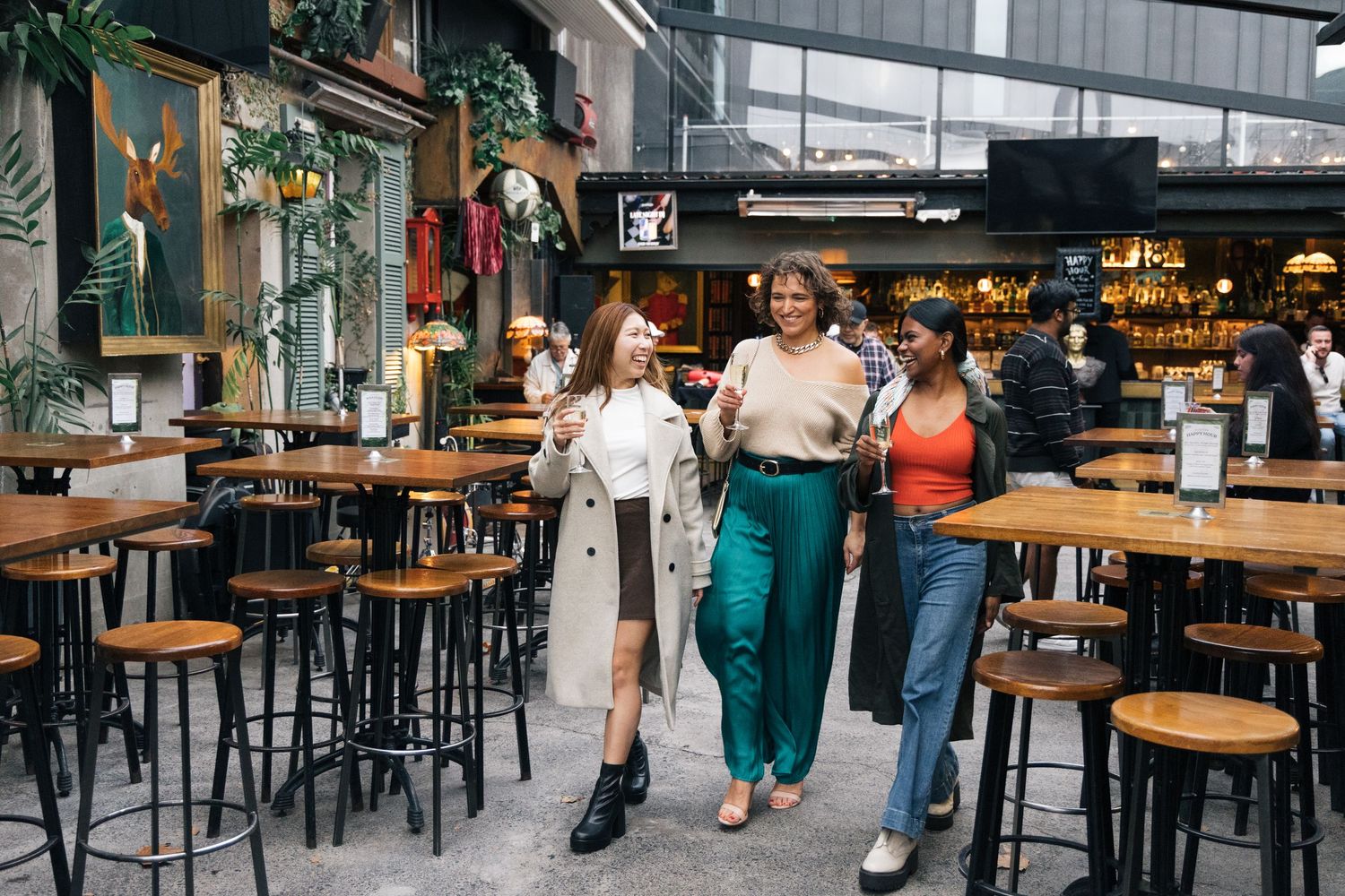 Three women walking and laughing together through a trendy indoor-outdoor bar venue.