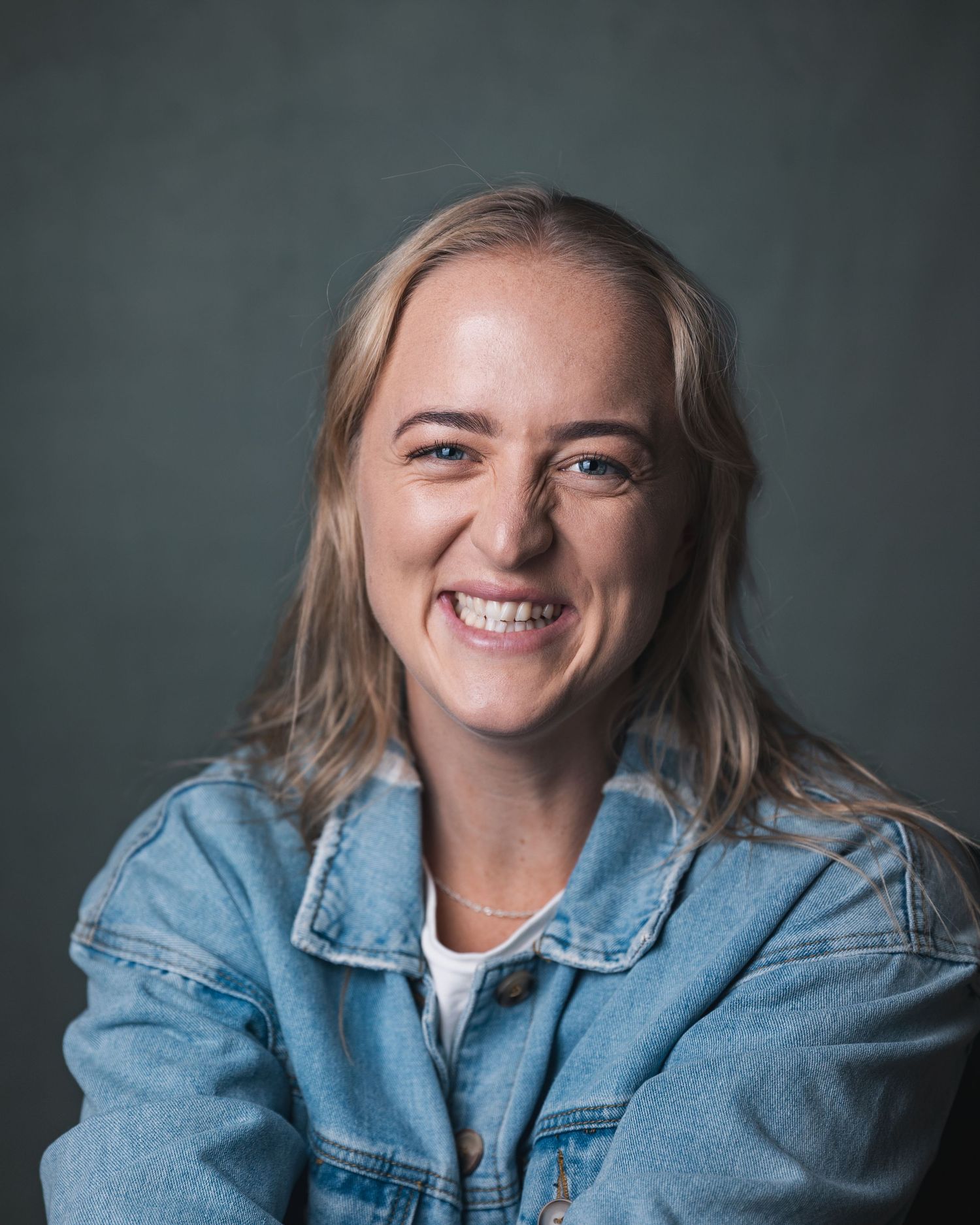 Smiling blonde woman in denim jacket against dark studio background in casual portrait.