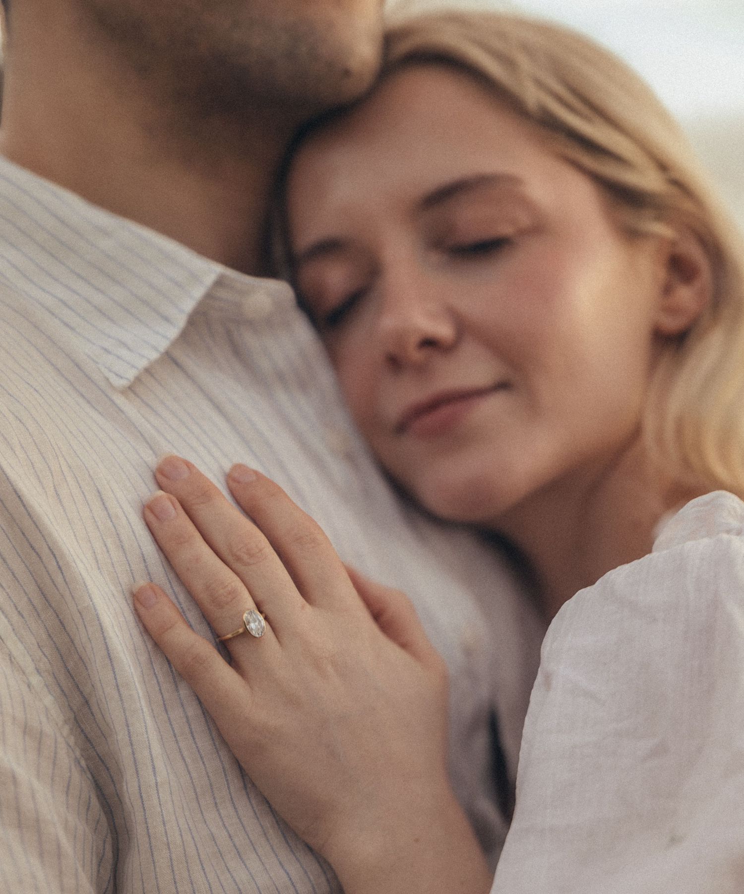 Woman with engagement ring rests her head on man's chest in a romantic, tender embrace.