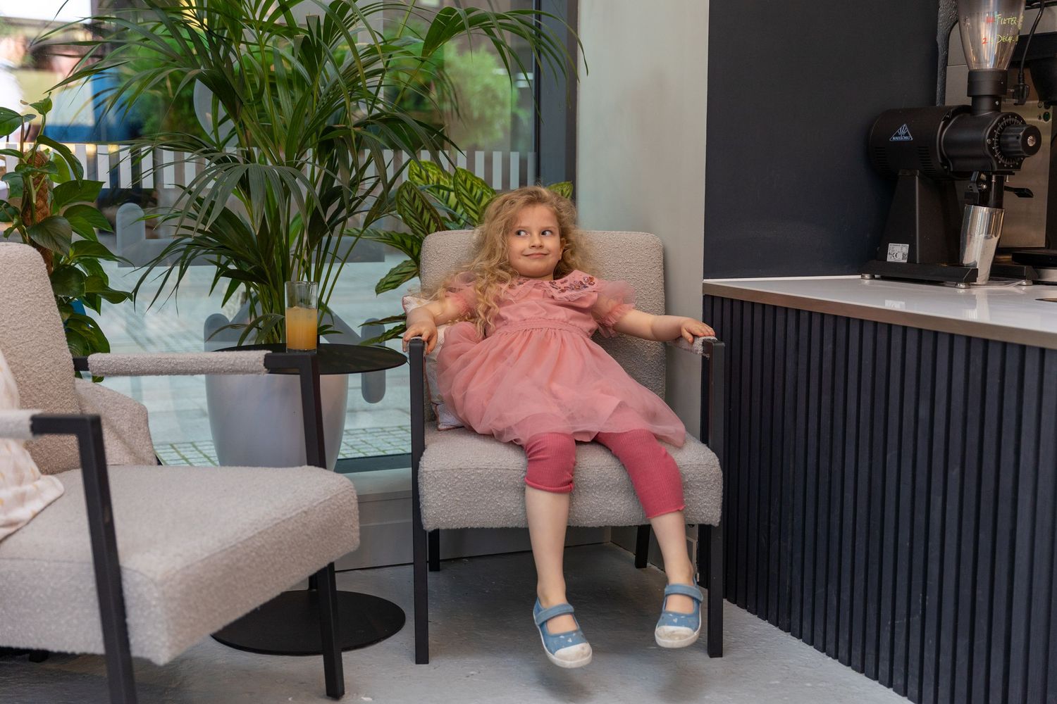 Young blonde girl relaxing in a grey chair at a modern cafe with indoor plants.