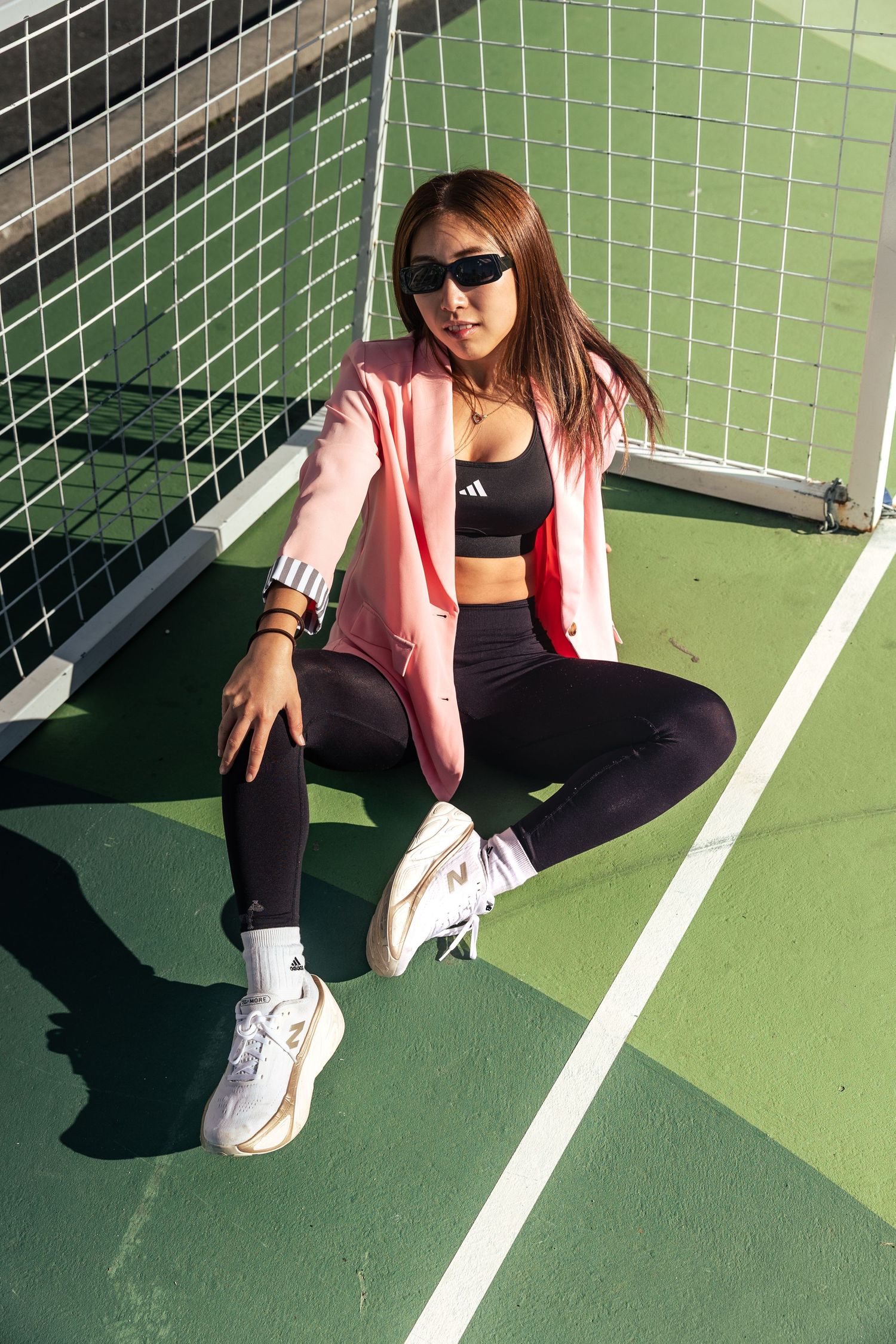 Stylish woman in pink blazer and black Adidas activewear sitting on a green tennis court.