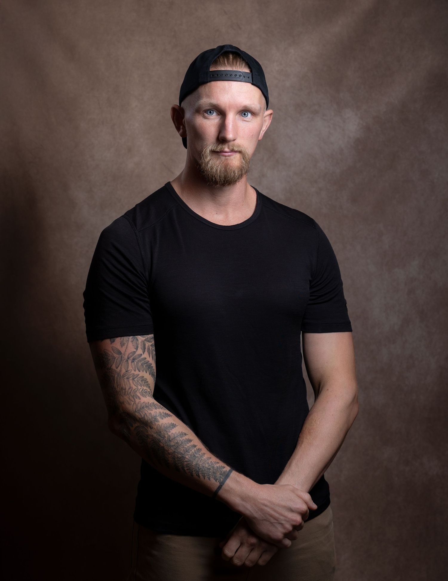Tattooed man in black t-shirt standing with hands clasped wearing backwards cap.