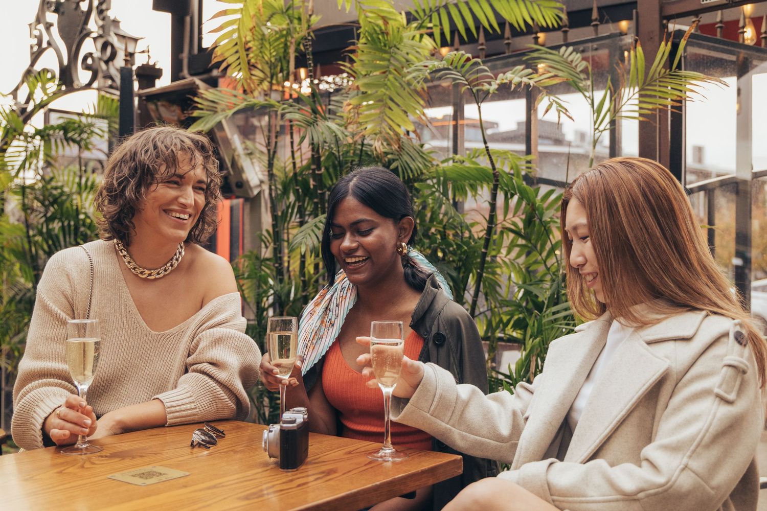 Three women laughing and toasting with champagne at an outdoor cafe table.