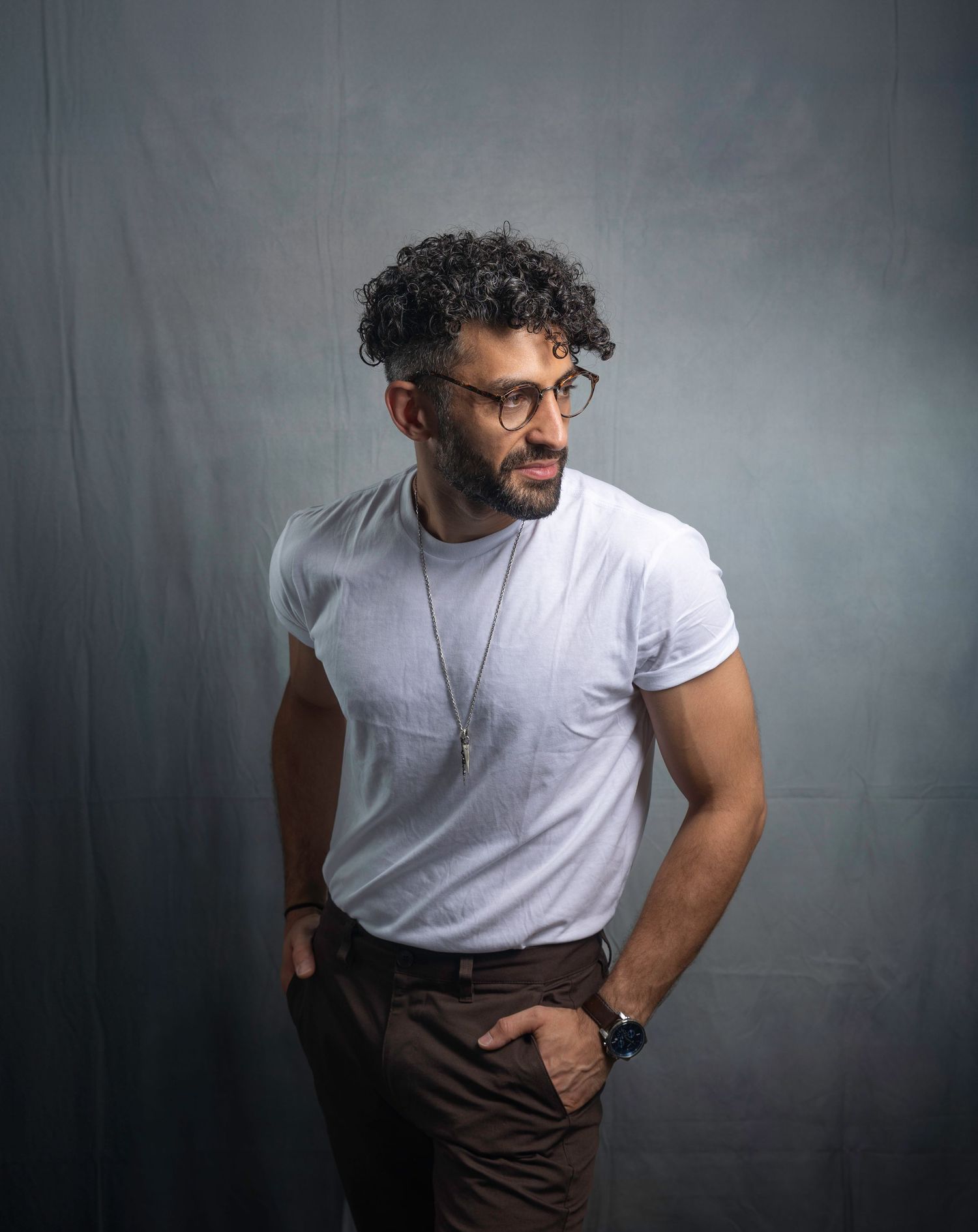 Muscular man with curly hair and glasses in white t-shirt posing against gray studio background.