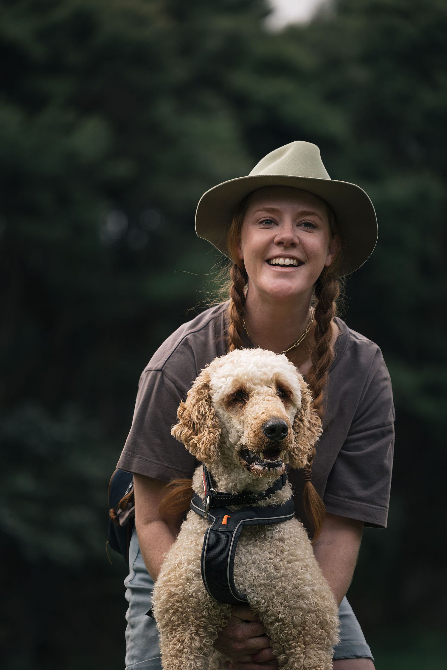 Smiling young woman in wide-brim hat hugging golden labradoodle dog outdoors with green trees behind.