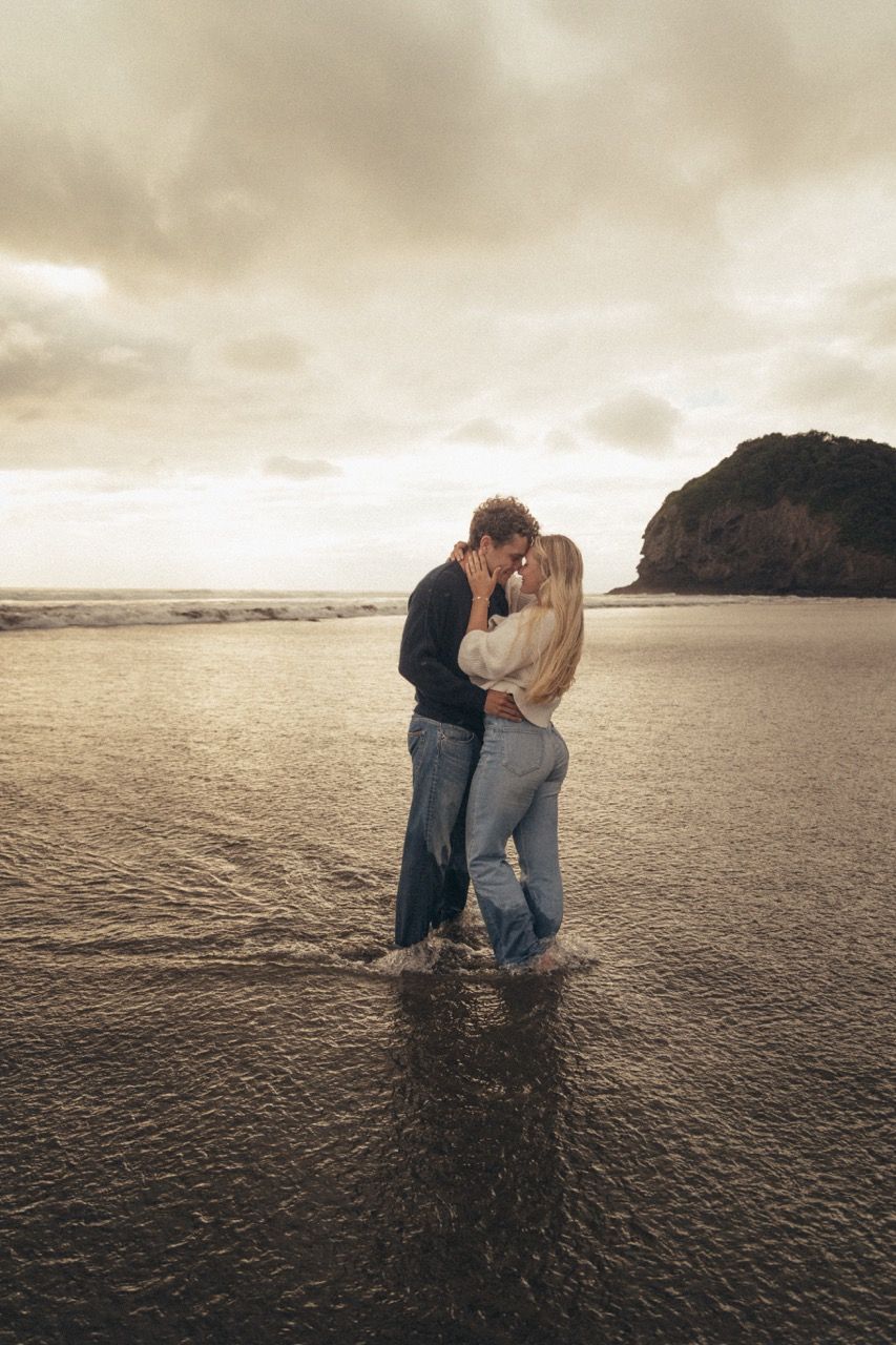 Couple kissing romantically on a wet beach at sunset with dramatic cloudy sky and rocky headland backdrop.