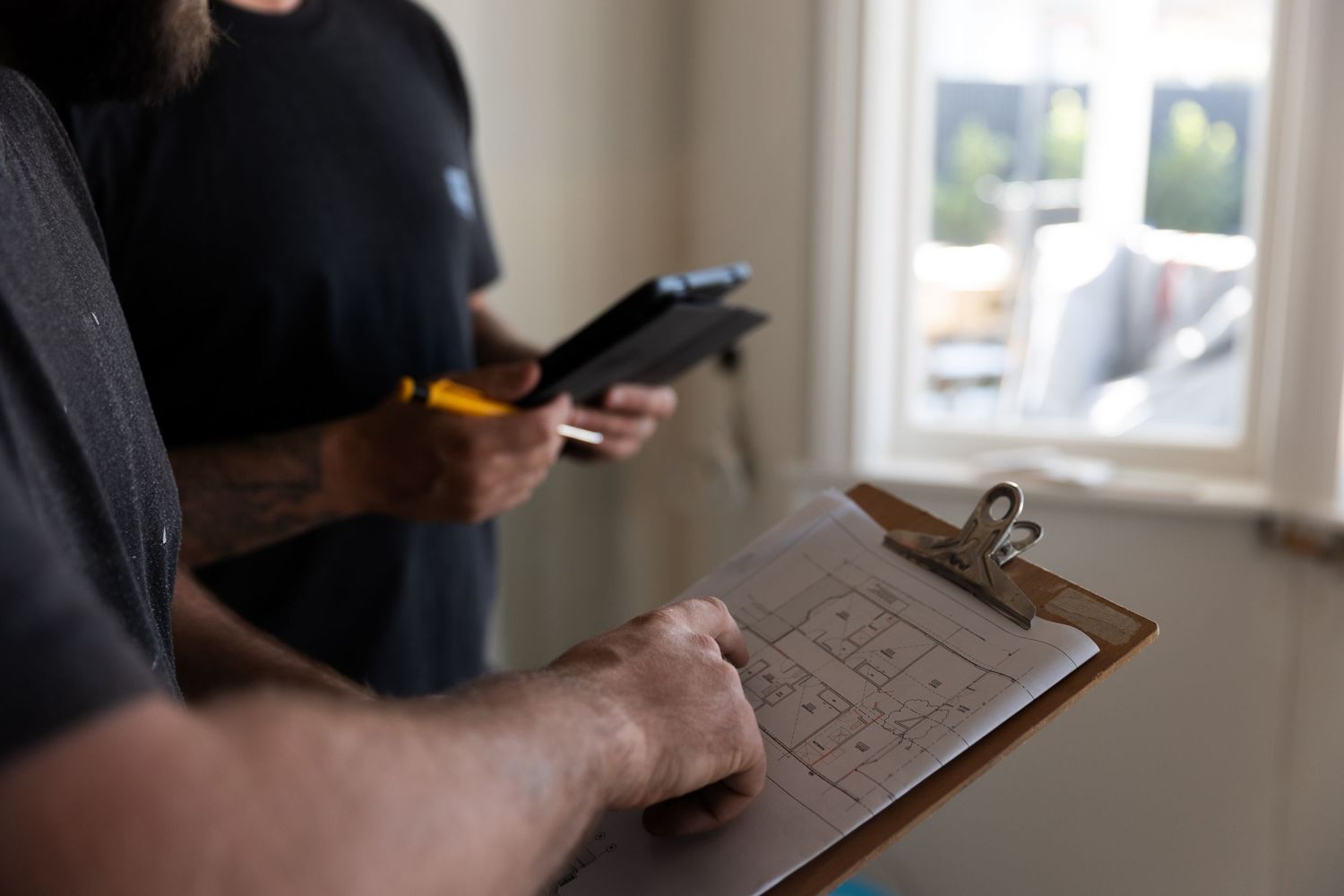 Two builders reviewing floor plan blueprints on a clipboard inside a construction site.