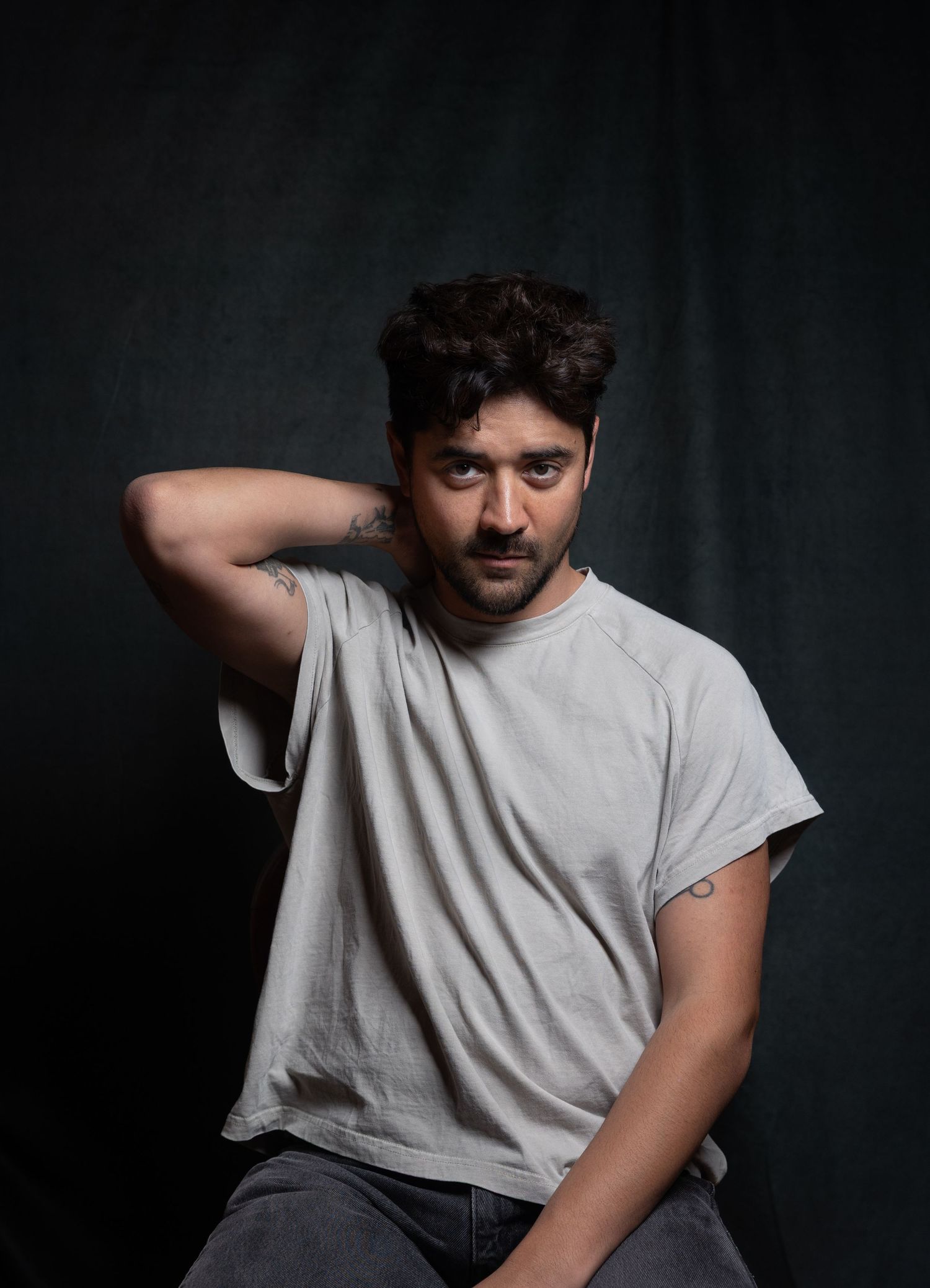 Young man in gray t-shirt posing with hand behind neck against dark background.