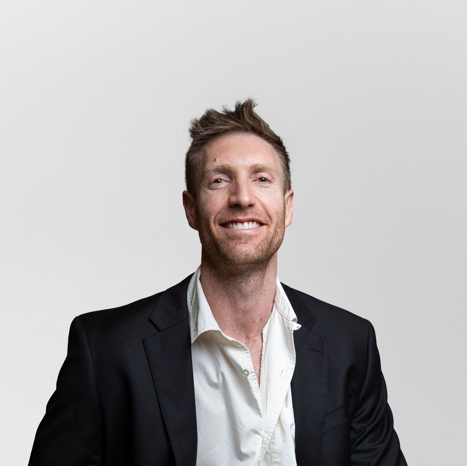 Smiling man in black blazer and white shirt posing against a light gray background.