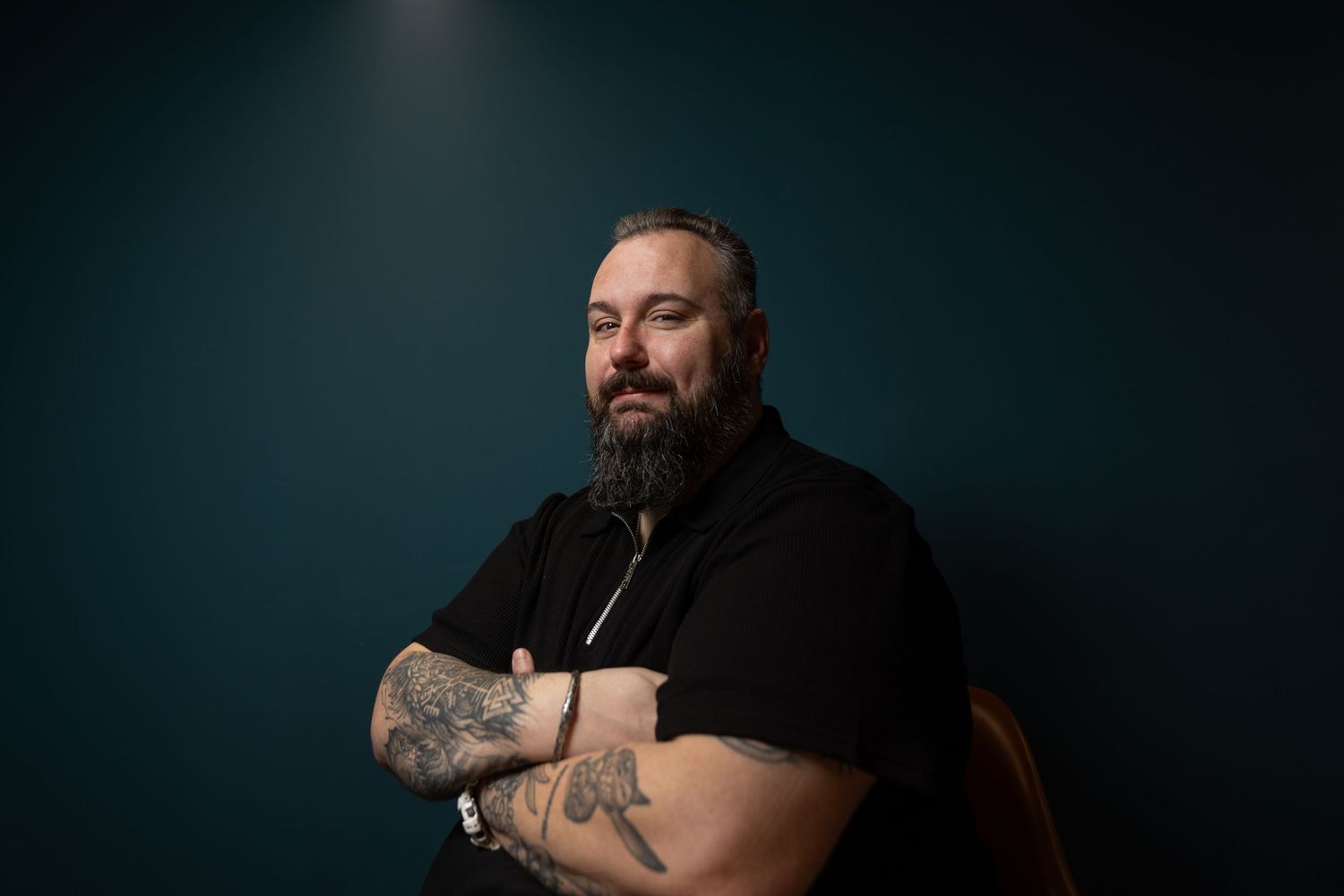 Tattooed bearded man in a black polo shirt with arms crossed against a dark teal background.