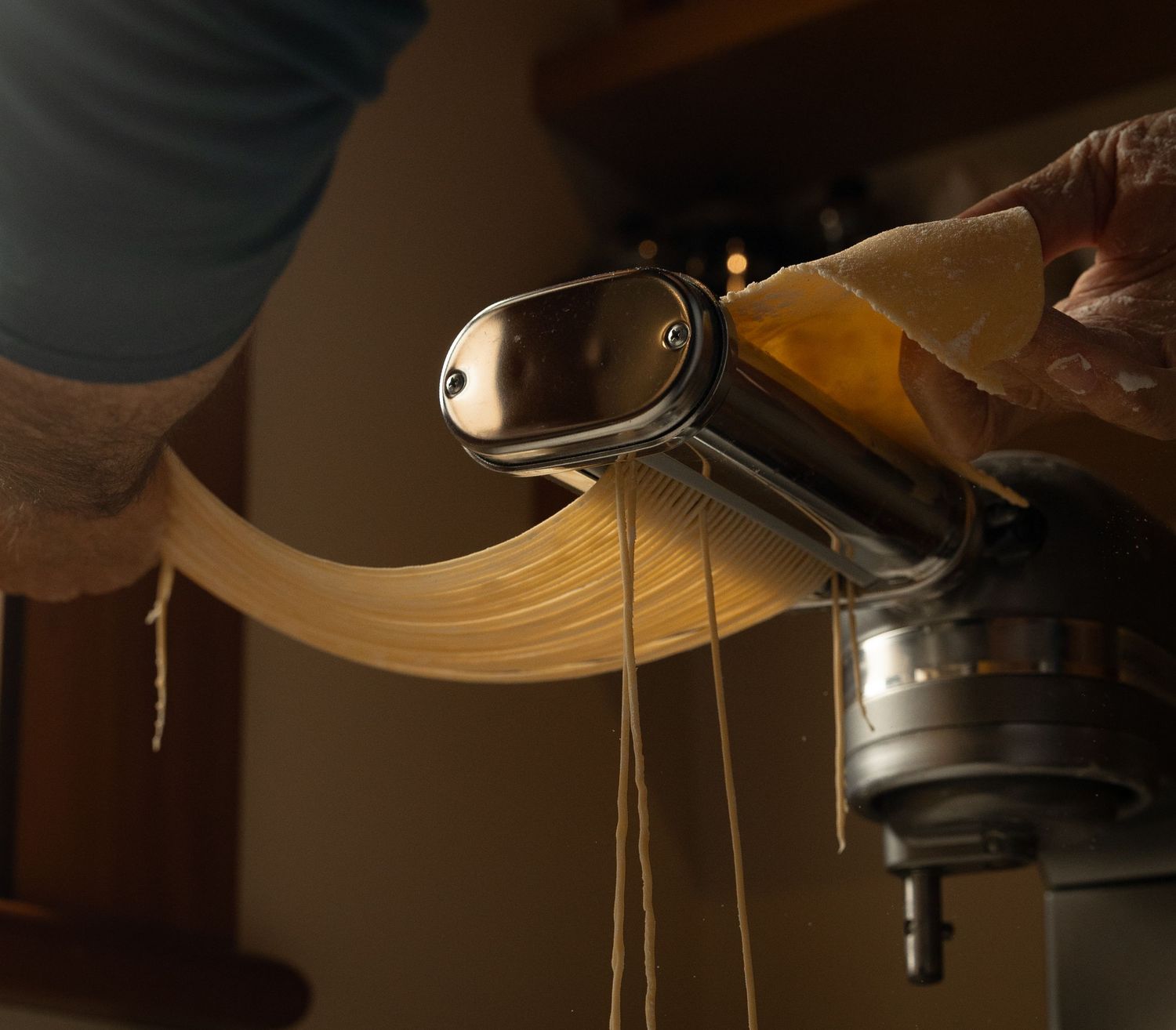 Fresh pasta sheets and strands feeding through a KitchenAid pasta cutter attachment.