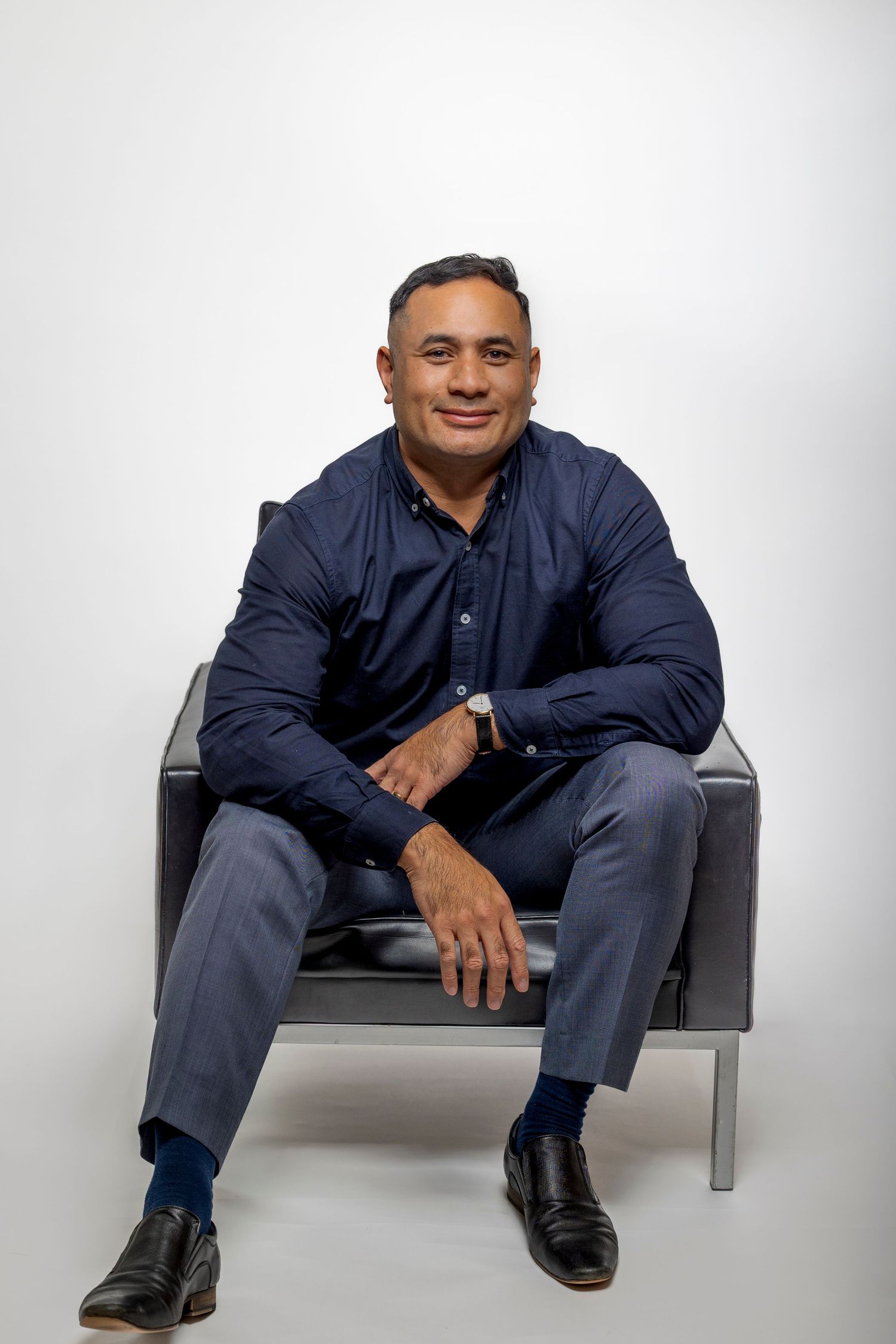 Smiling man in navy blue shirt sitting casually in a leather chair on white background.