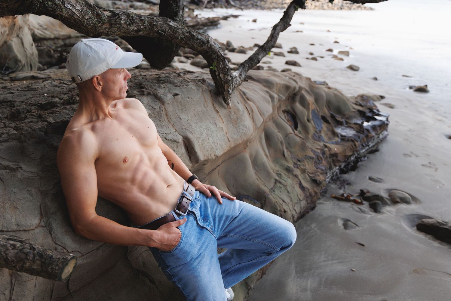 Muscular shirtless man in white cap and jeans relaxing on rocky beach near driftwood.
