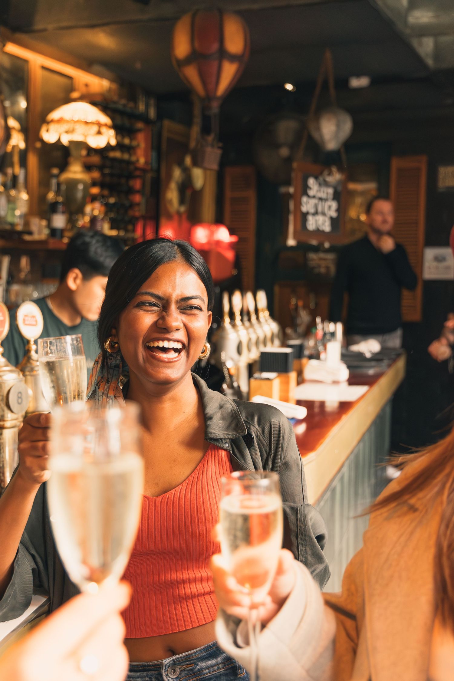 A laughing woman toasts with champagne glasses with friends at a warm, bustling bar.