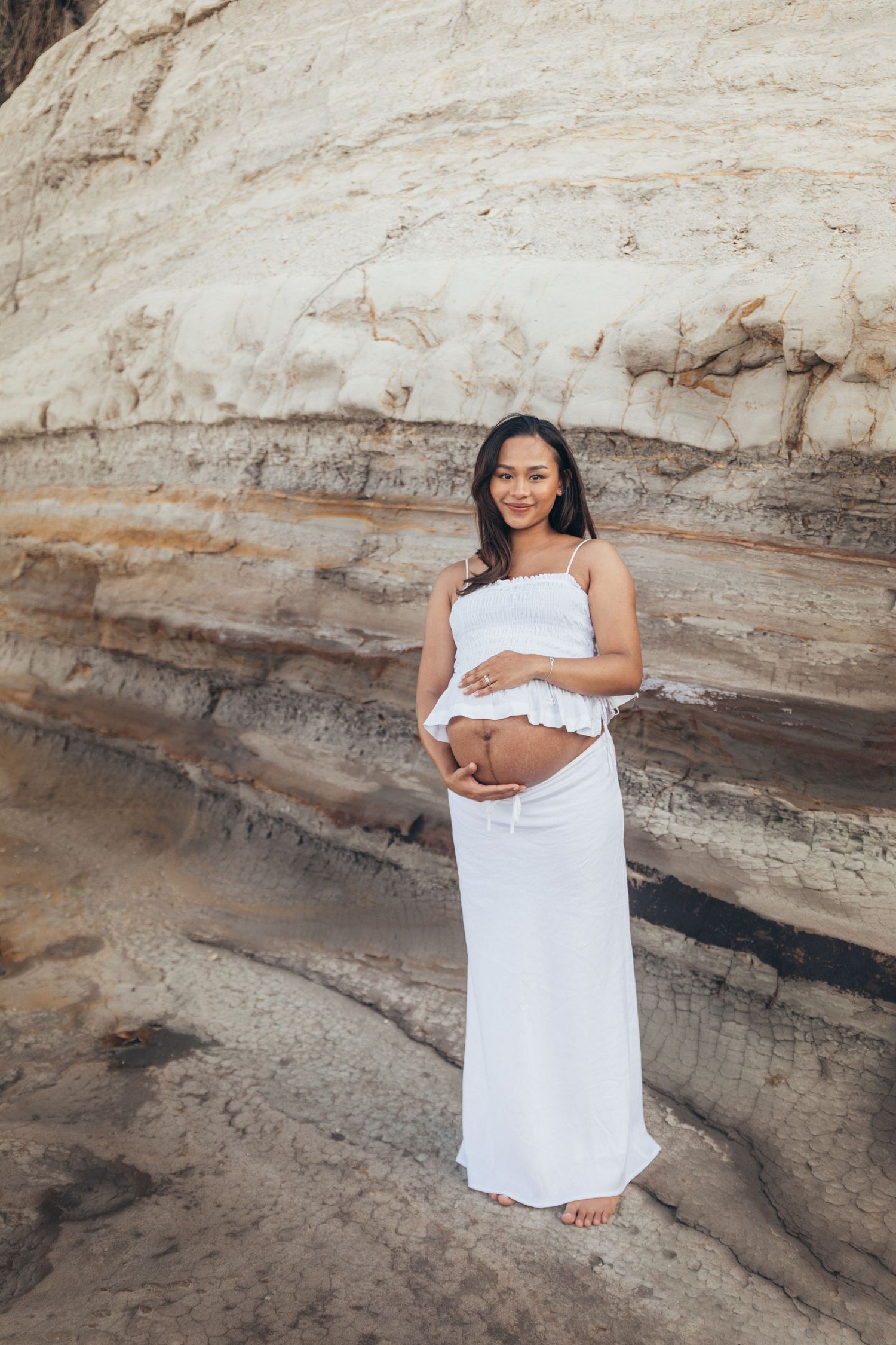 Pregnant woman in white dress cradling baby bump against sandy rock formation backdrop.