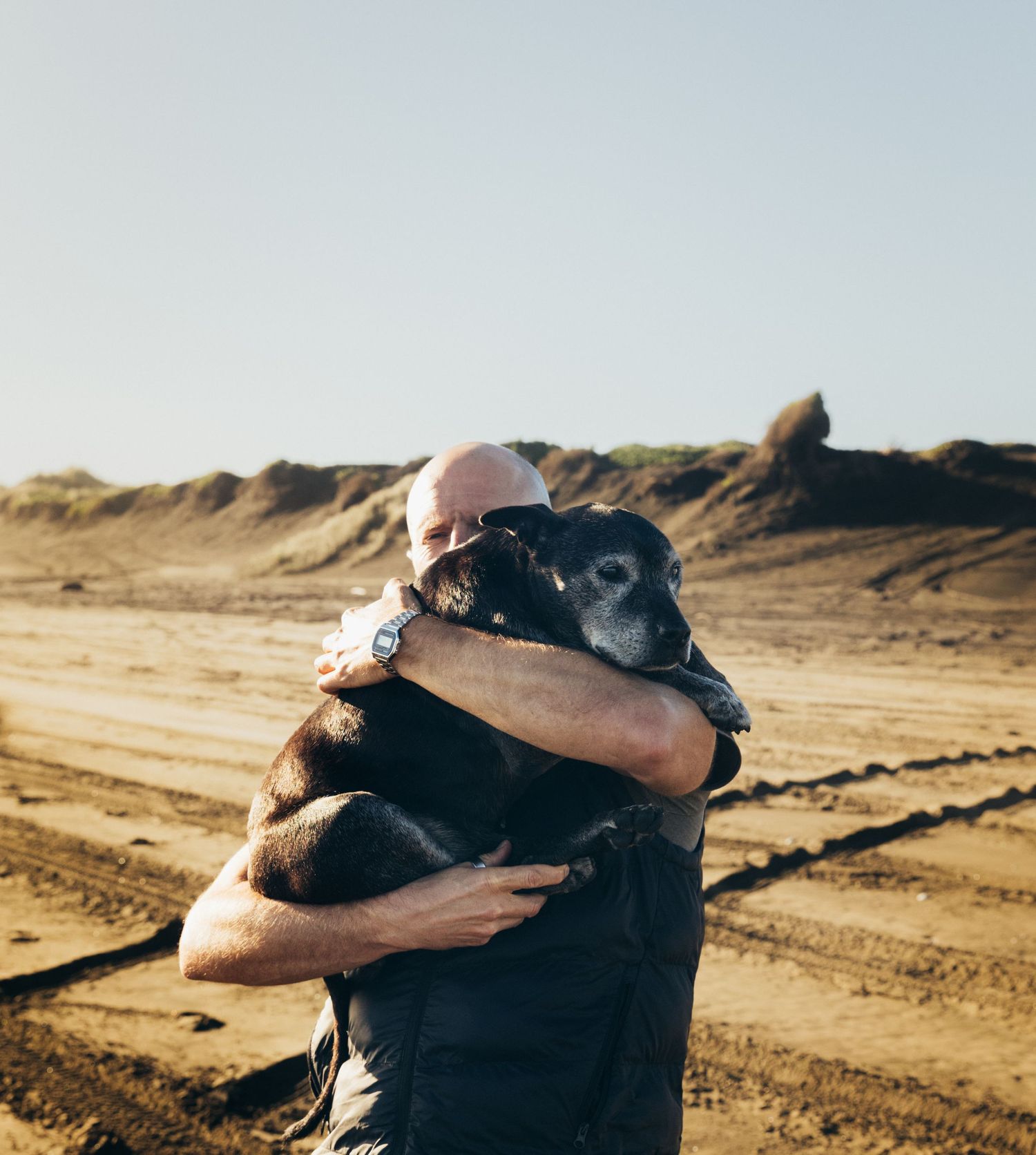 Bald man tenderly hugs his black senior dog outdoors in a sandy landscape.