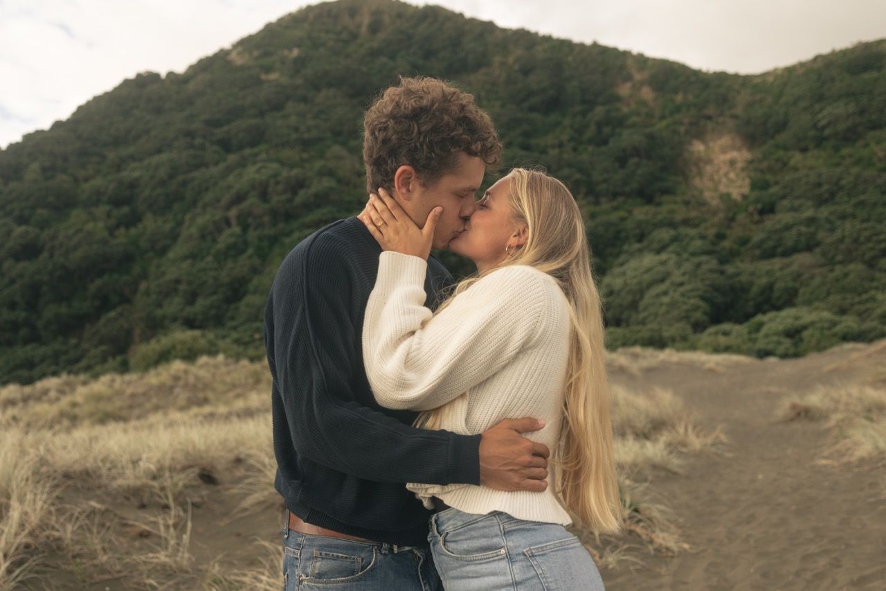 Couple sharing a romantic kiss outdoors with lush green mountain landscape in the background.
