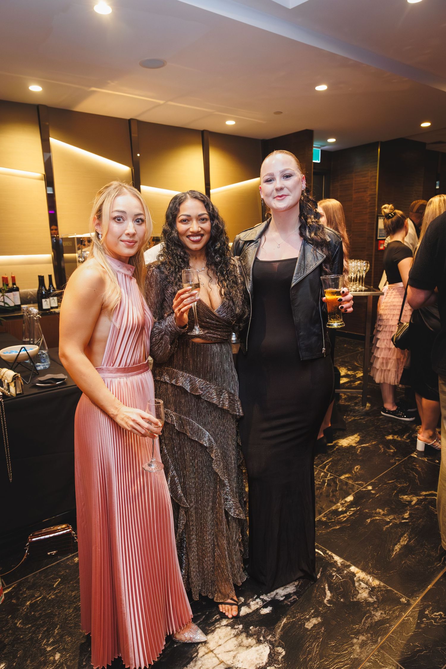 Three elegantly dressed women pose together at a glamorous evening event, holding champagne glasses.