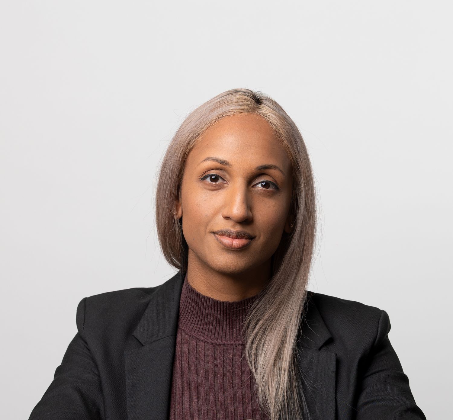 Professional headshot of a young woman with long blonde highlighted hair wearing a black blazer and burgundy turtleneck.
