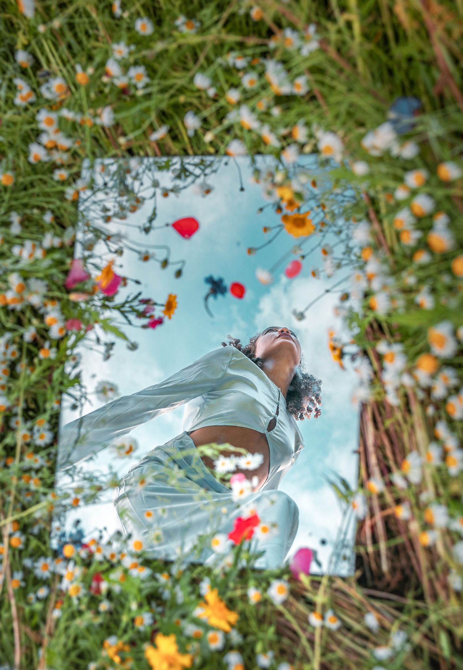 Mirror reflection of a woman in white outfit surrounded by colorful wildflowers including daisies and poppies.