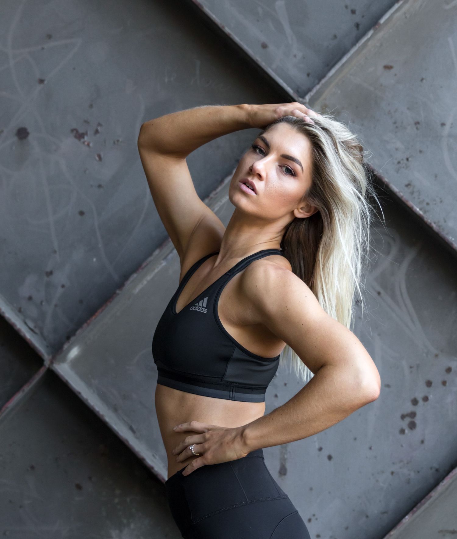 Fit blonde woman in black Adidas sports bra posing against a geometric metal panel wall.