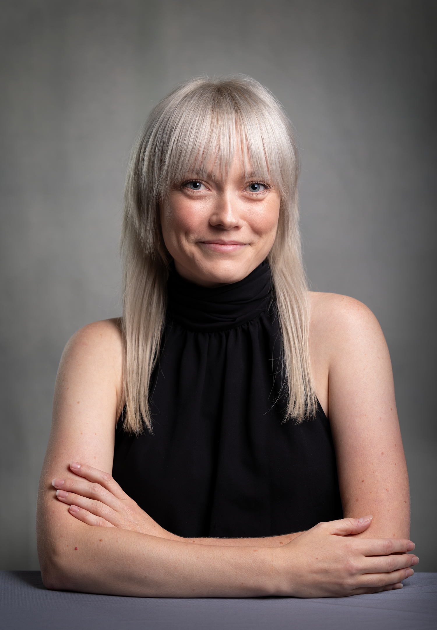 Smiling blonde woman with bangs wearing black sleeveless turtleneck, arms crossed, studio portrait.