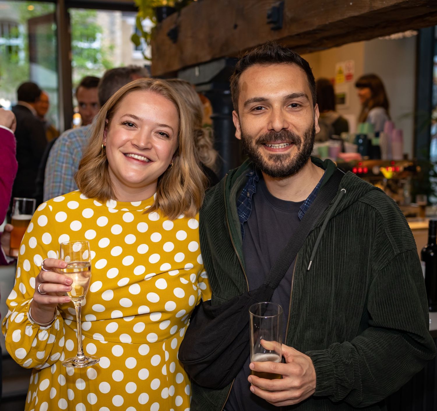A smiling woman in a yellow polka dot dress and bearded man enjoy drinks at a social event.