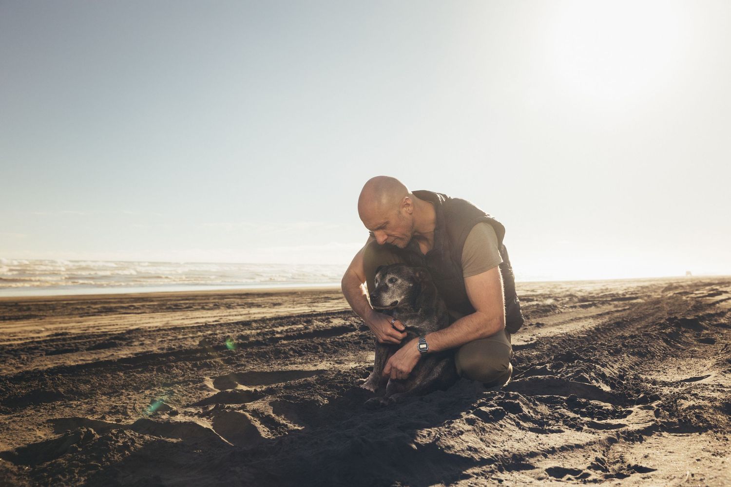 Bald man kneeling on sandy beach tenderly petting his small black dog in warm sunlight.