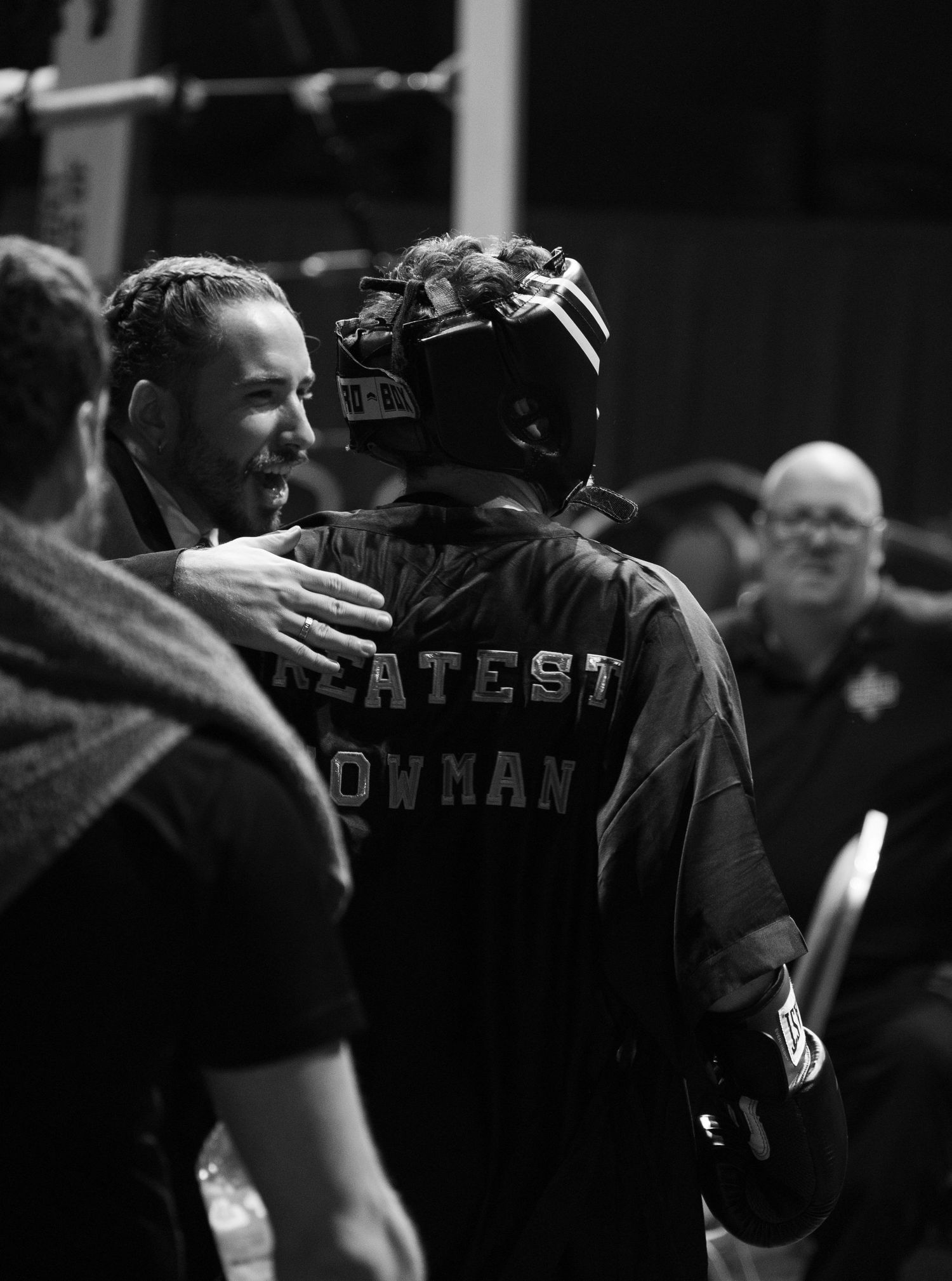 Two men embrace near a boxing ring, one wearing a Greatest Showman jacket and helmet.