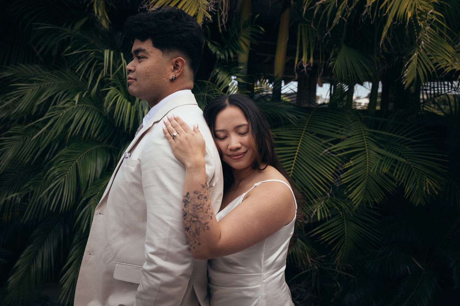 A couple in white wedding attire poses romantically among lush tropical palm leaves outdoors.