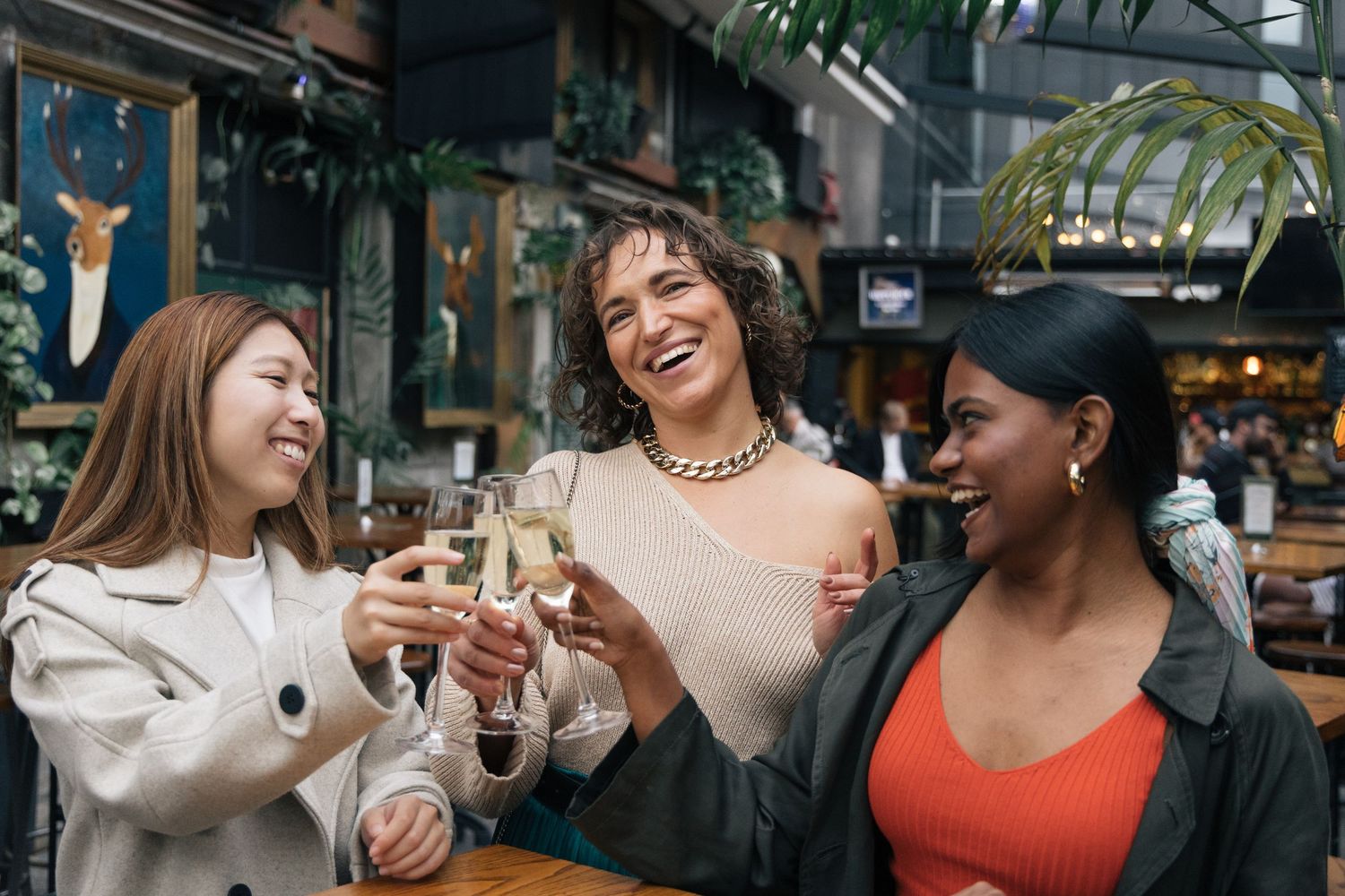 Three diverse women laughing and toasting with champagne glasses at a stylish indoor bar or restaurant.