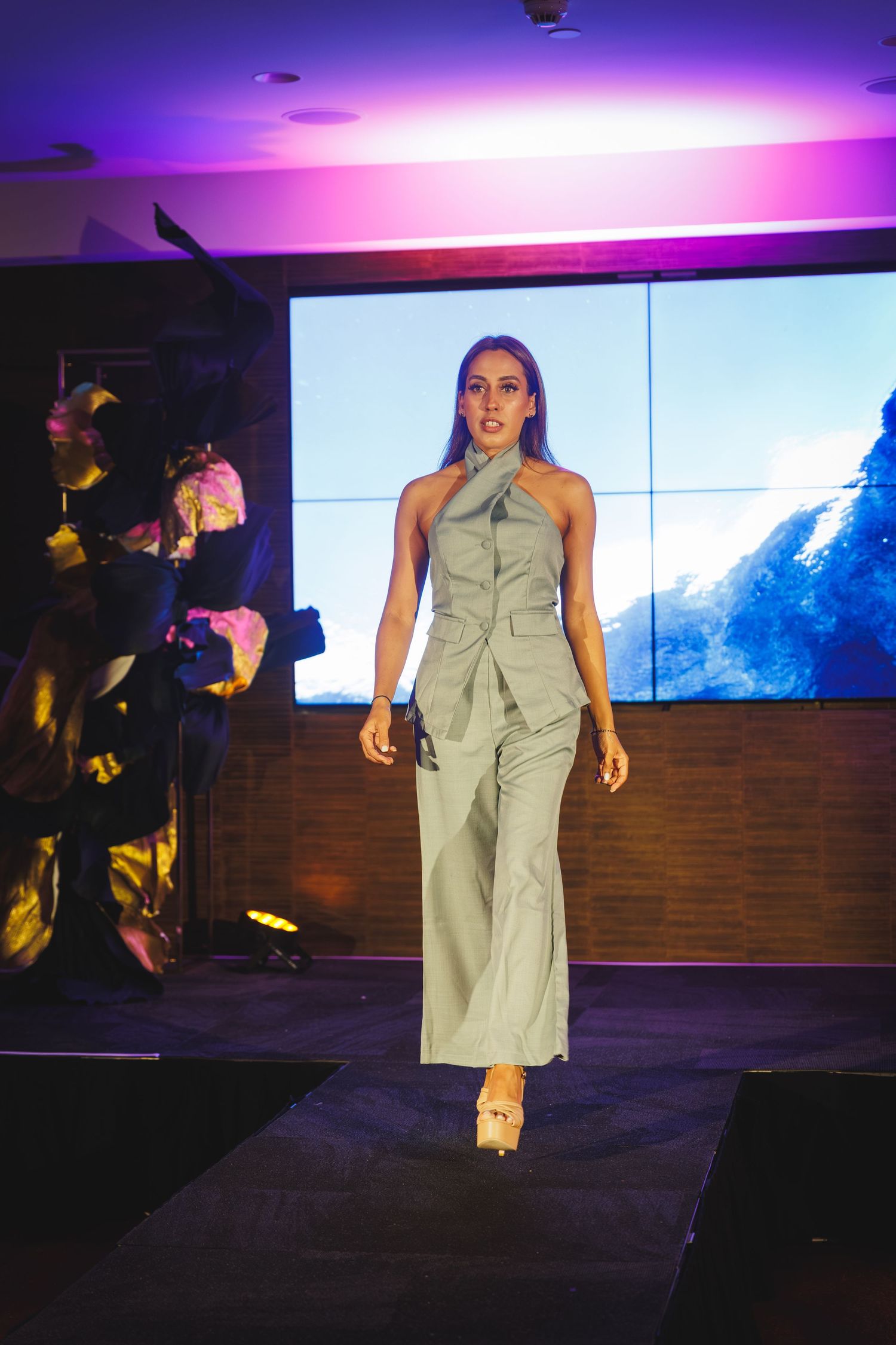 Female model walks the runway in a sage green halter jumpsuit at a fashion show with purple lighting.