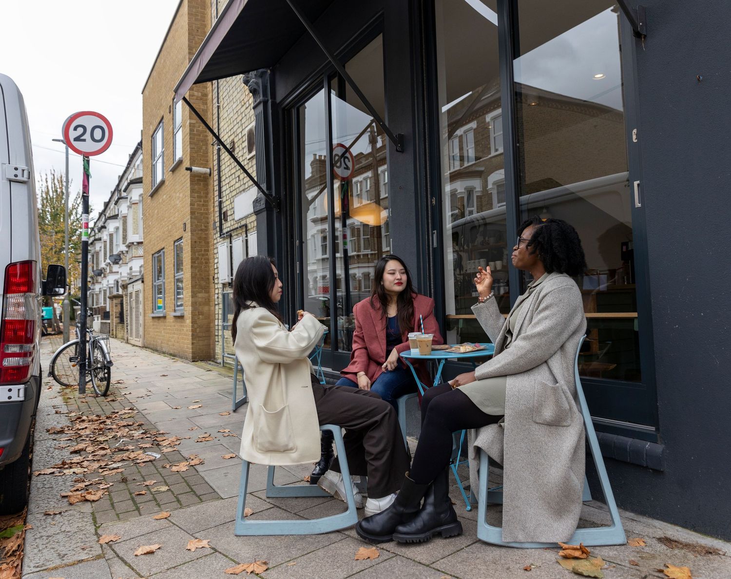 Three women sitting outdoors at a cafe table on a London street beside a 20mph speed limit sign.