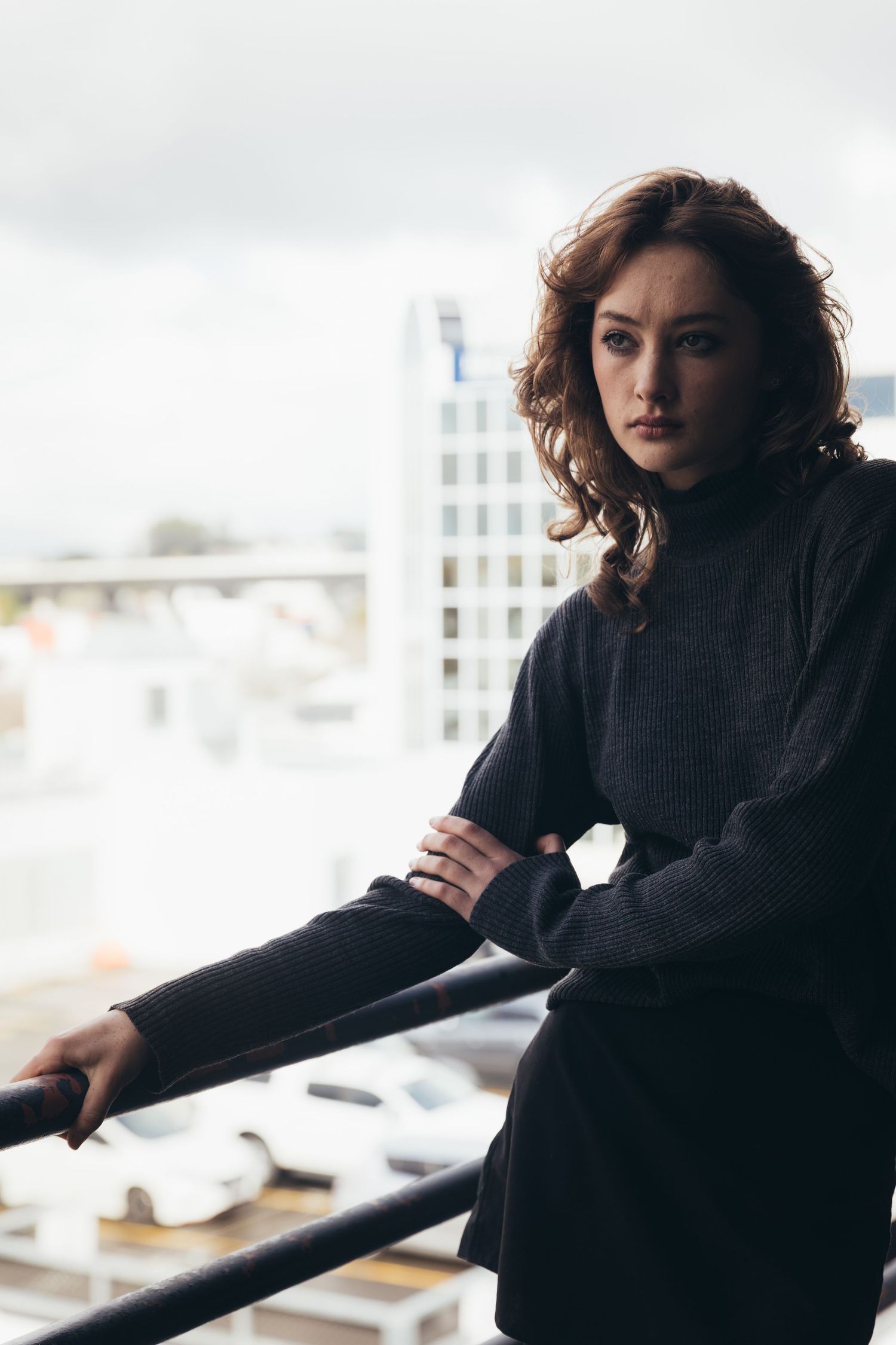 Young woman with curly auburn hair in dark sweater leaning on railing with city skyline behind her.