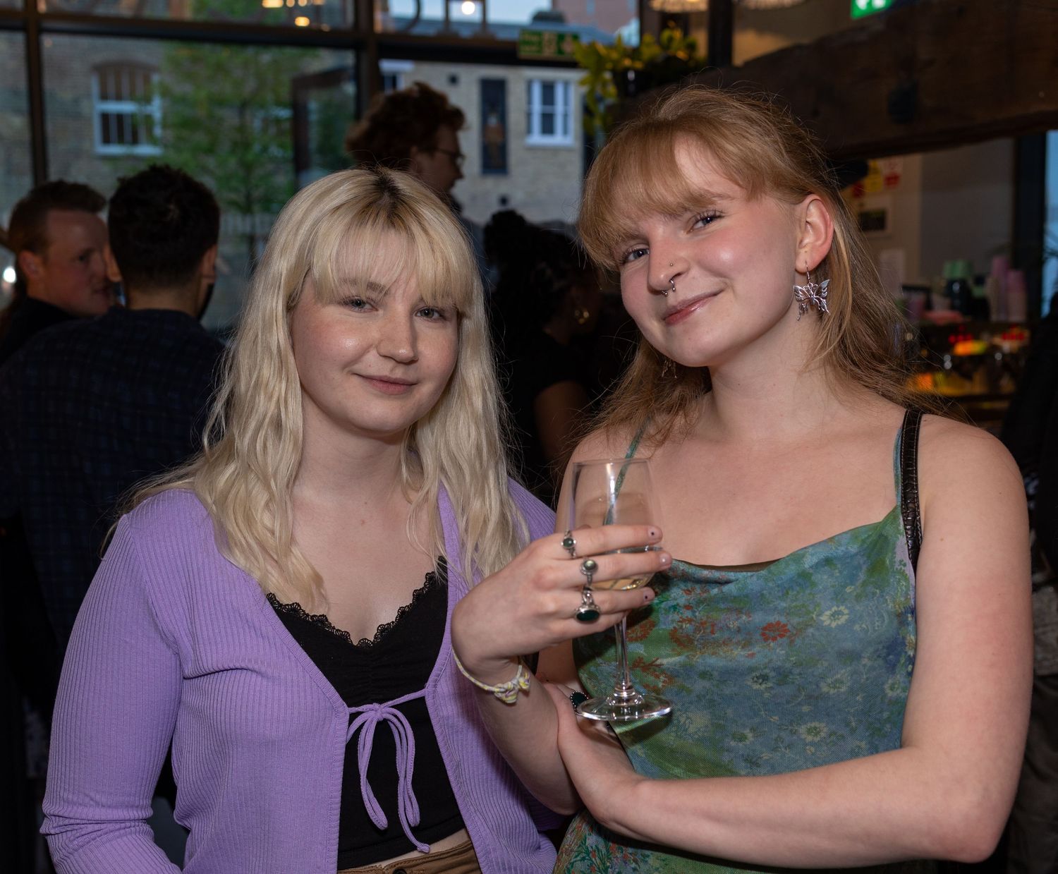 Two young blonde women smile at a social event, one holding a glass of wine.