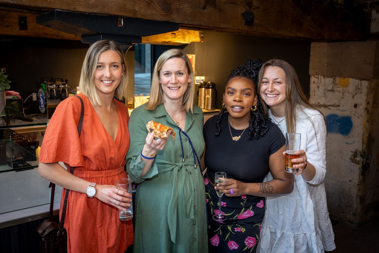 Four women smile together holding drinks and food at a casual indoor party.
