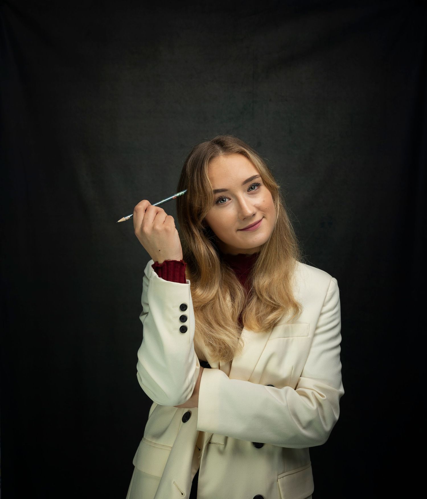 Young blonde woman in white blazer holding pen thoughtfully in a professional studio portrait.
