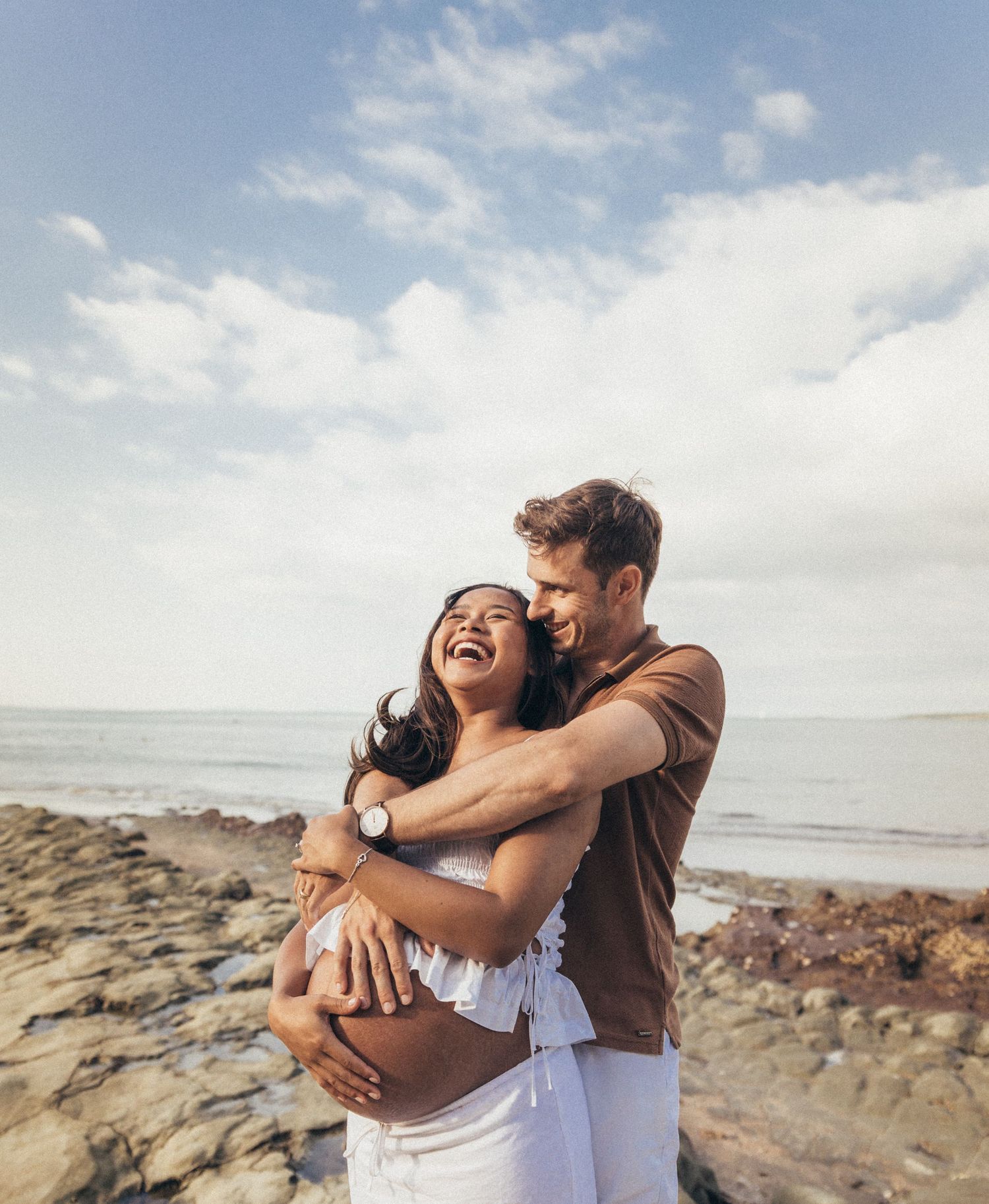 Happy pregnant couple embracing on rocky beach, laughing together under a partly cloudy sky.