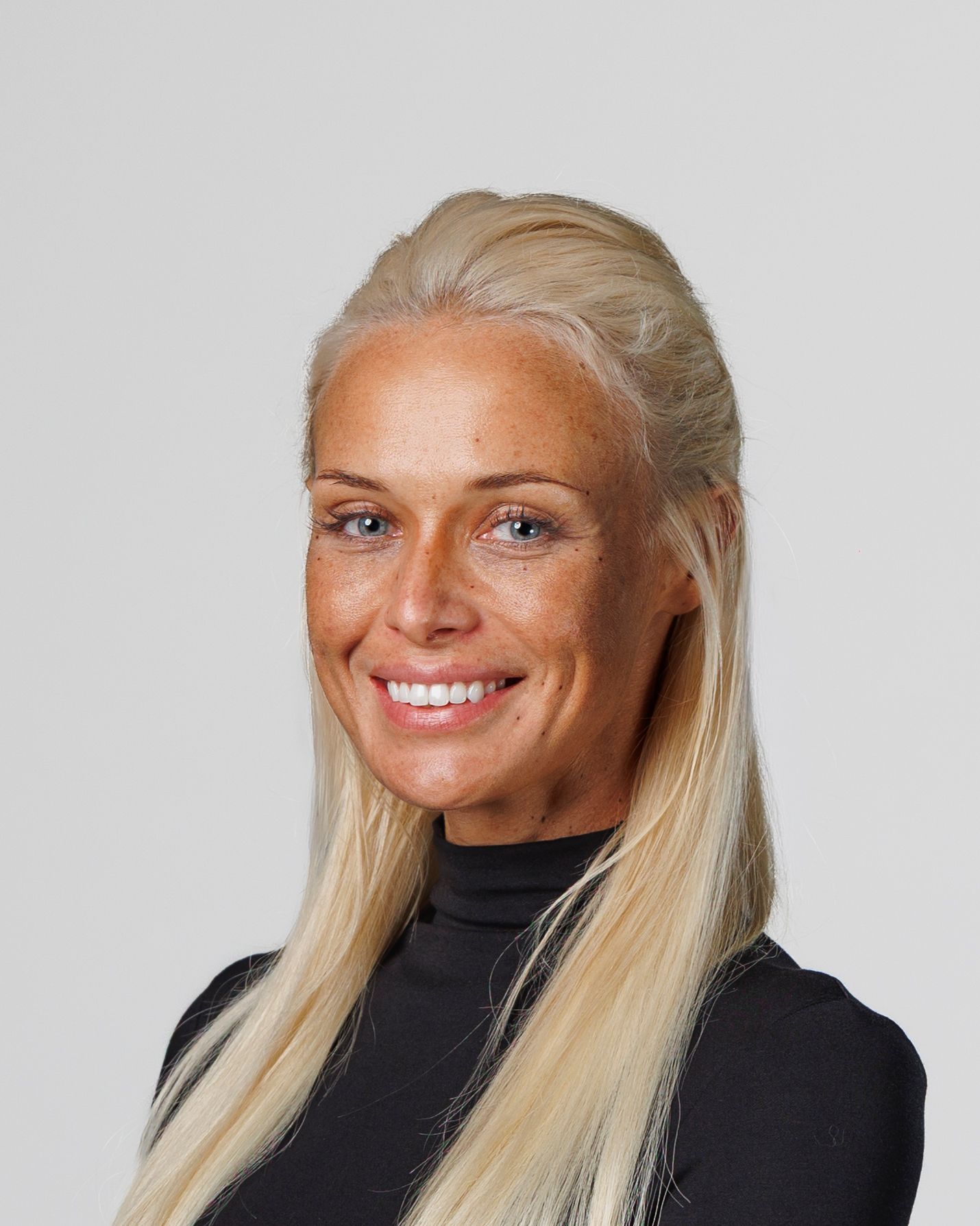 Smiling woman with long blonde hair and freckles wearing a black turtleneck against a gray background.