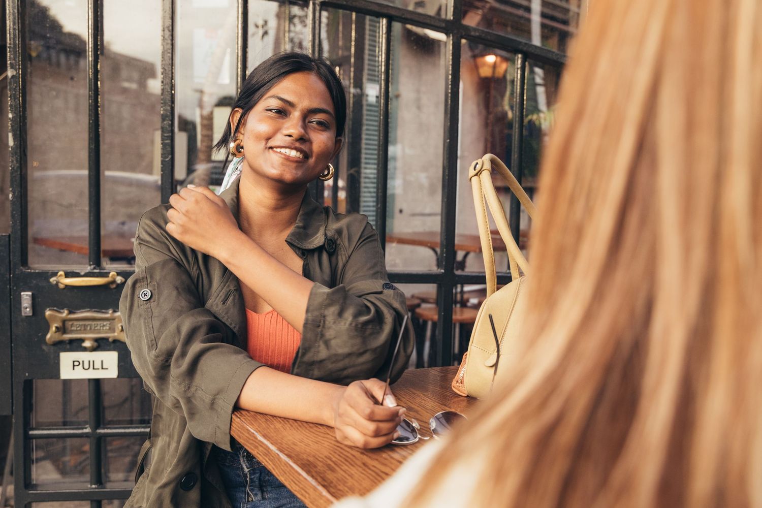Two women smiling and chatting at an outdoor cafe table, one wearing a green jacket and hoop earrings.