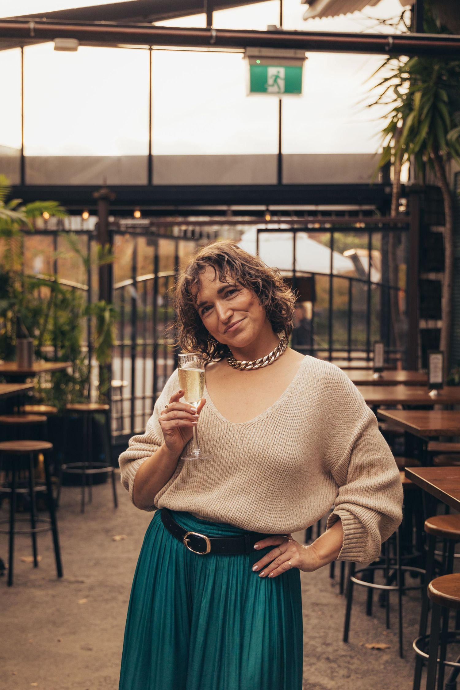 Woman in teal skirt and beige sweater holding champagne glass at outdoor restaurant patio.