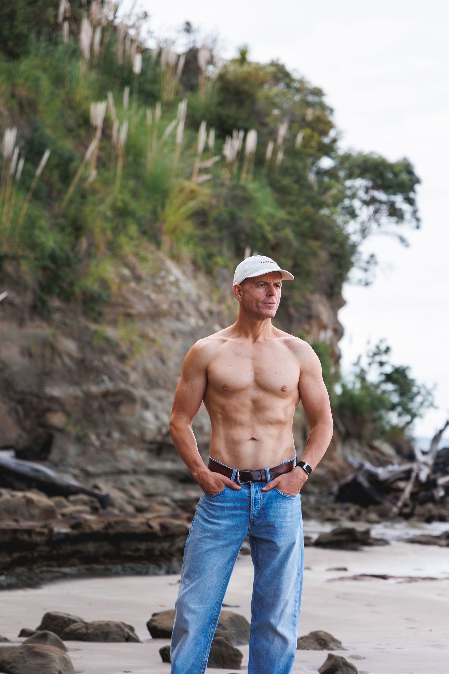 Muscular shirtless man in jeans and white cap standing on a rocky beach with cliffs.