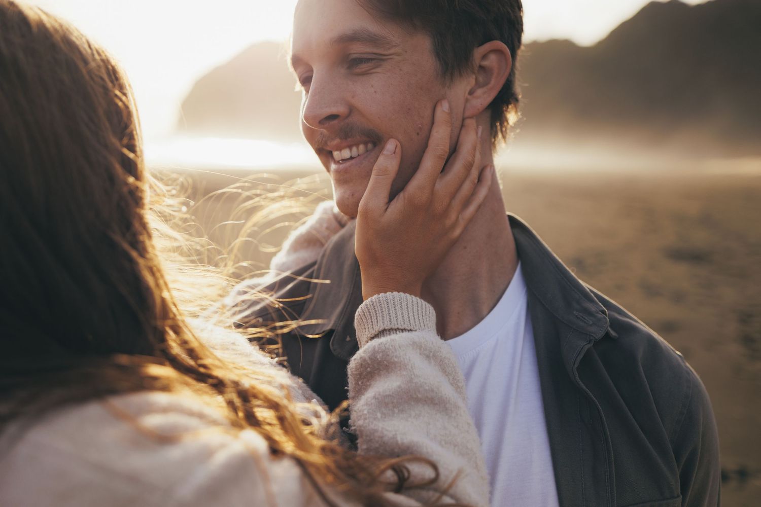 Romantic couple on a beach at sunset, woman tenderly touching smiling mans face with wind-blown hair.