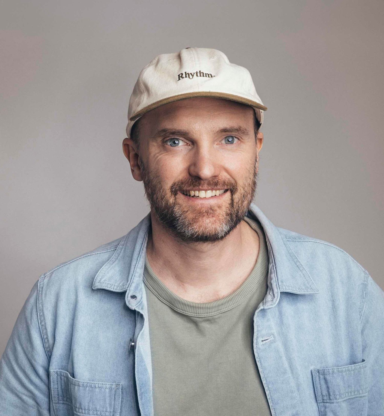 Smiling man with beard wearing a cream Rhythm cap and blue denim shirt against a gray background.