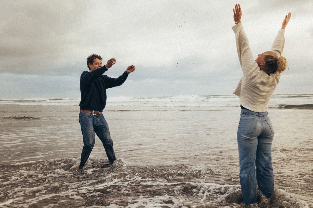 Couple playing joyfully on a cloudy beach, splashing water and raising arms in the air near ocean waves.