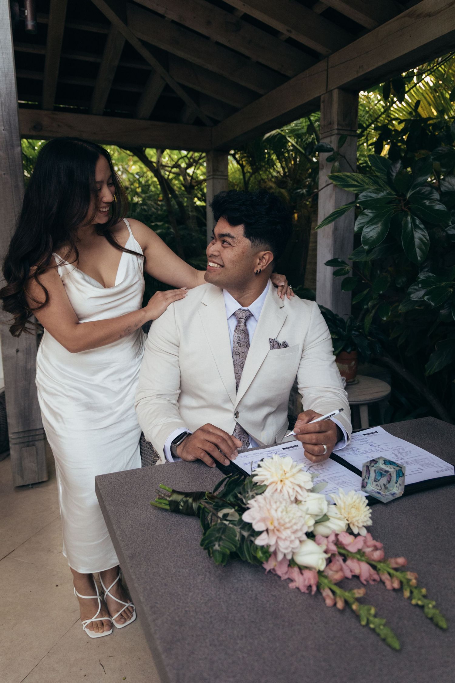 Bride in white dress and groom in cream suit signing wedding documents at outdoor ceremony table with floral bouquet.