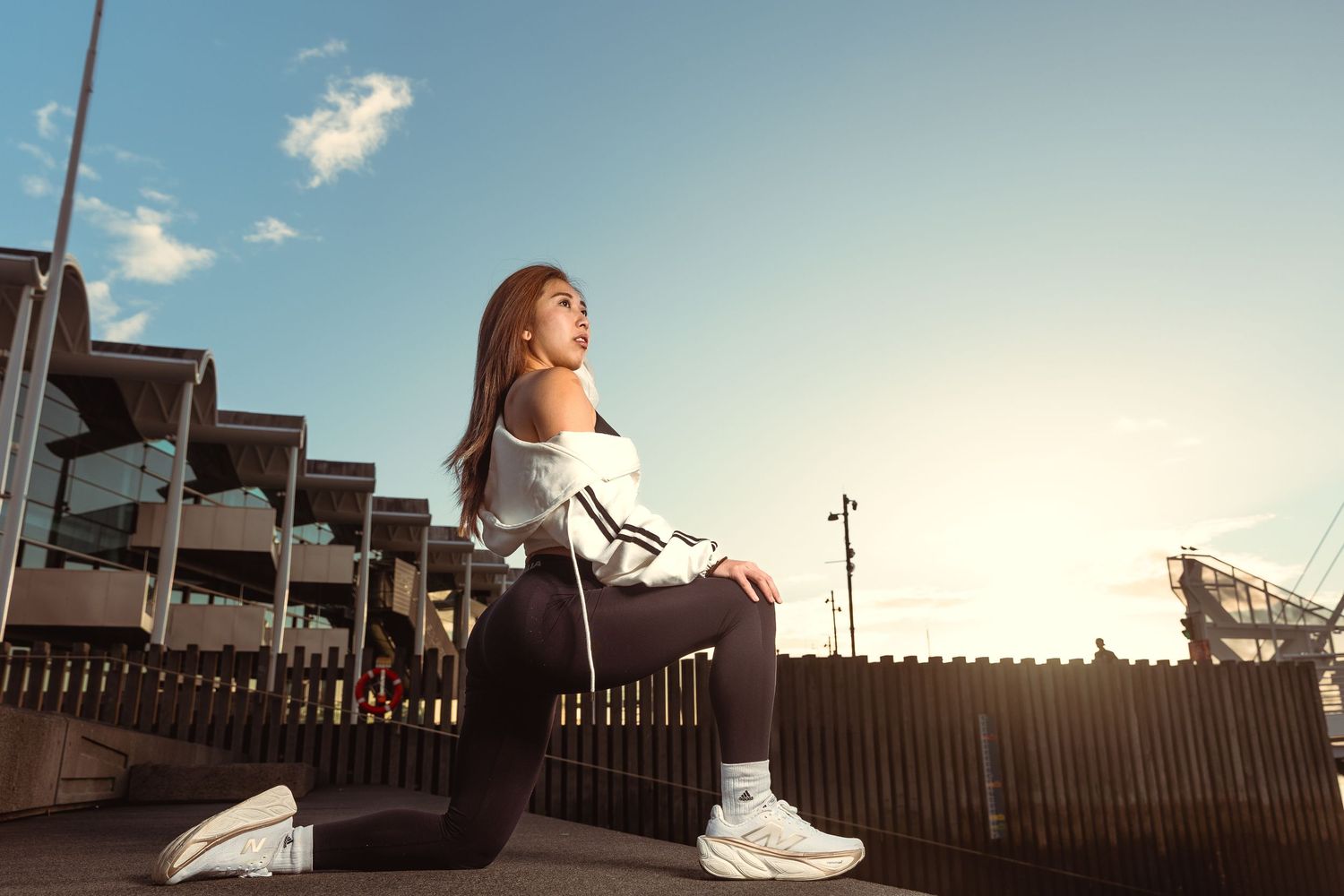 Young woman in white jacket and black leggings sitting outdoors at sunset in an urban setting.