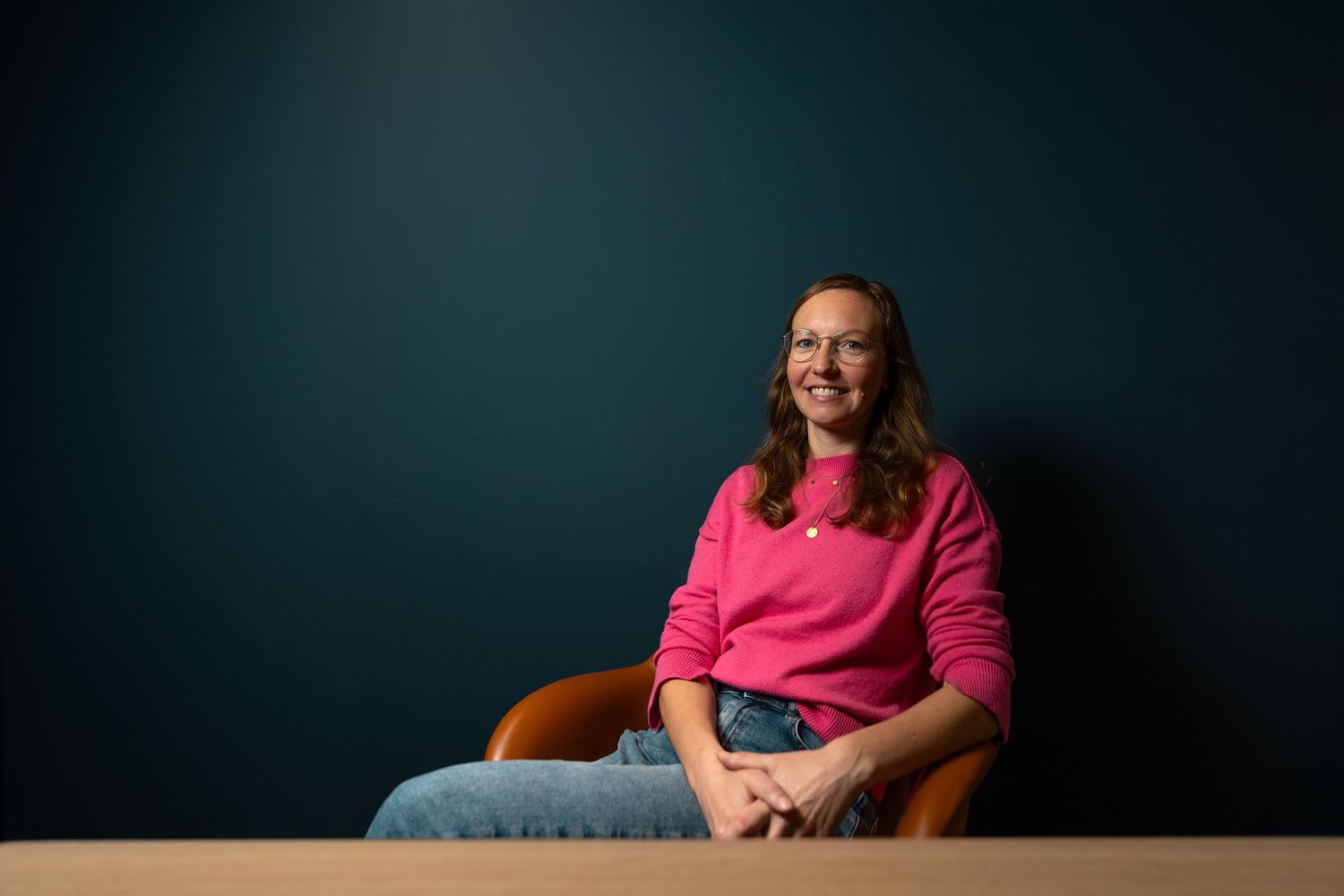 Smiling woman in a bright pink sweater and jeans seated at a table against a dark teal background.