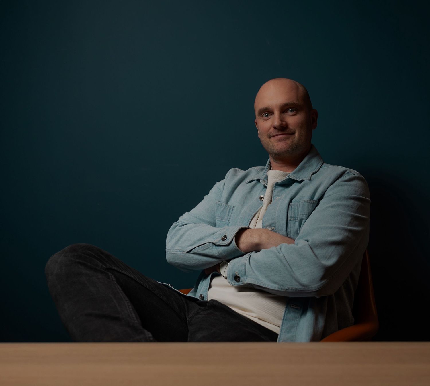 Bald man sitting relaxed with arms crossed wearing a light blue shirt against a teal wall.